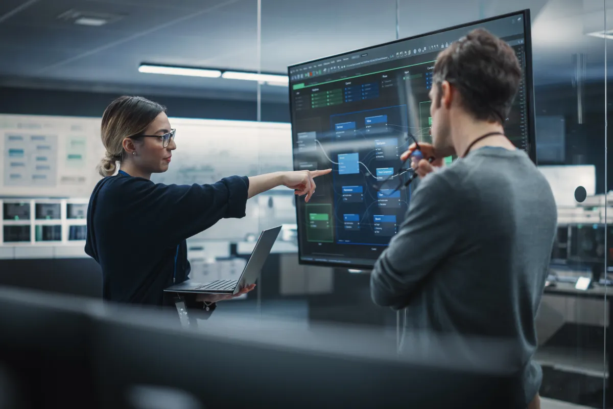 A woman holding a laptop points to a complex software diagram on a large screen as she discusses it with a man in a modern office.