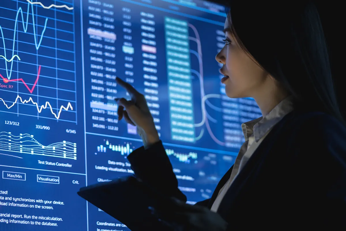 A woman points at a large, glowing blue screen displaying complex graphs and data in a dark room.