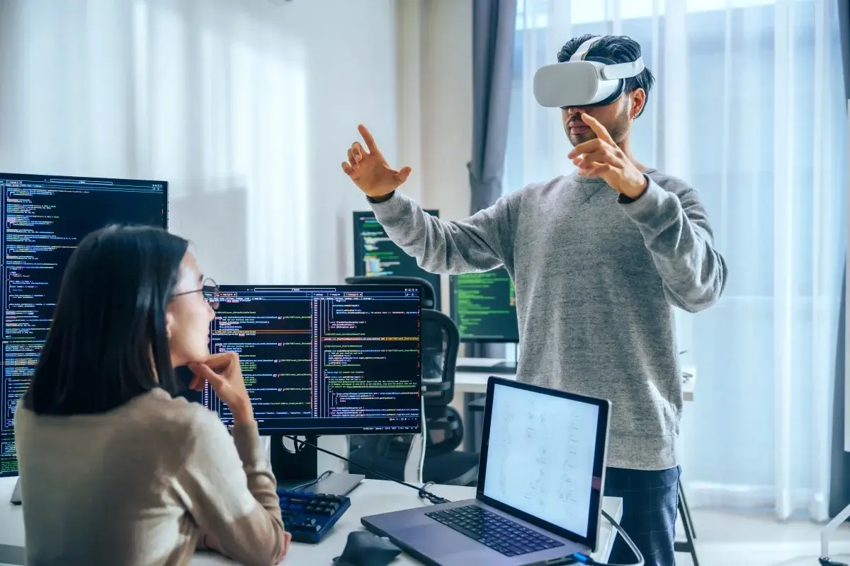 Asian software developer wearing a virtual reality headset works on a VR project, with a colleague focused on her computer
